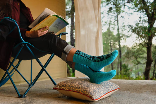 A person sitting in a chair with their feet up on a cushion, wearing handknitted motif panel socks in blue and green colors.