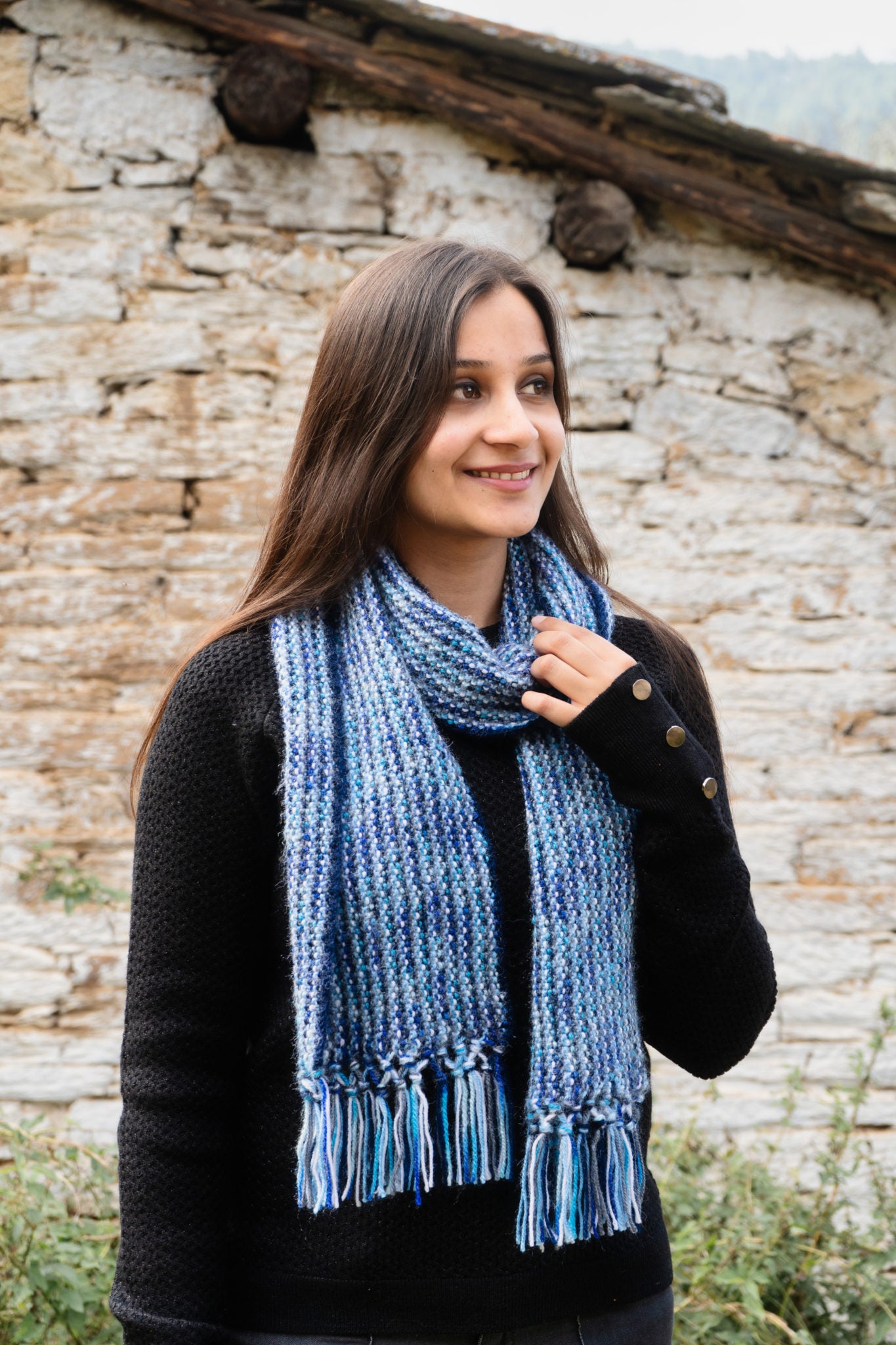 A woman standing outdoors wearing a black top and blue-striped long scarf with fringes.