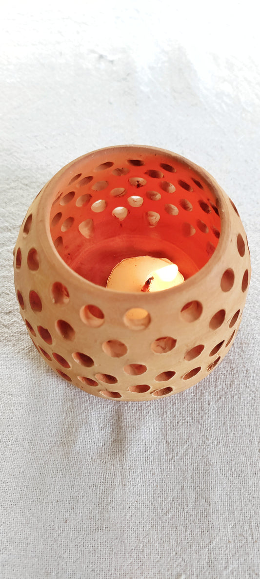 A beige terracotta tealight candle holder with a round shape and perforated design, containing a lit candle, placed on a white surface.